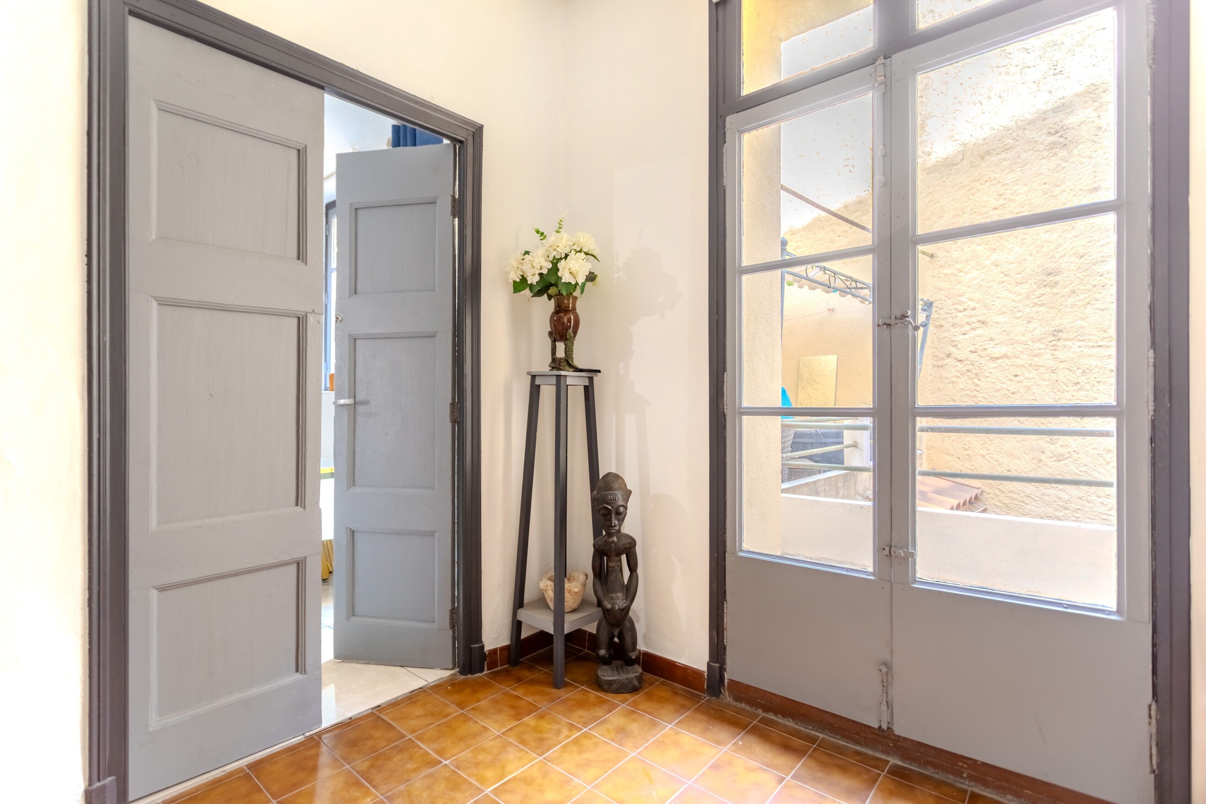 A hallway with a tiled floor, a white door, a window with a view of a sandy area, and a small table with a vase of flowers.