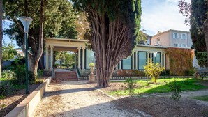 Un sentier de jardin menant à un bâtiment orné de colonnes, surmonté d’une pergola et bordé d’un grand arbre.