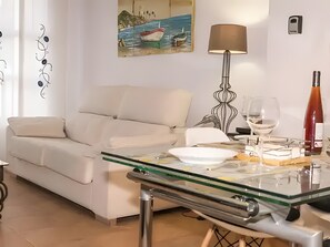 Un salon avec une table basse en verre, un canapé blanc, un tableau représentant un bateau accroché au mur, une lampe sur pied et une bouteille de vin.