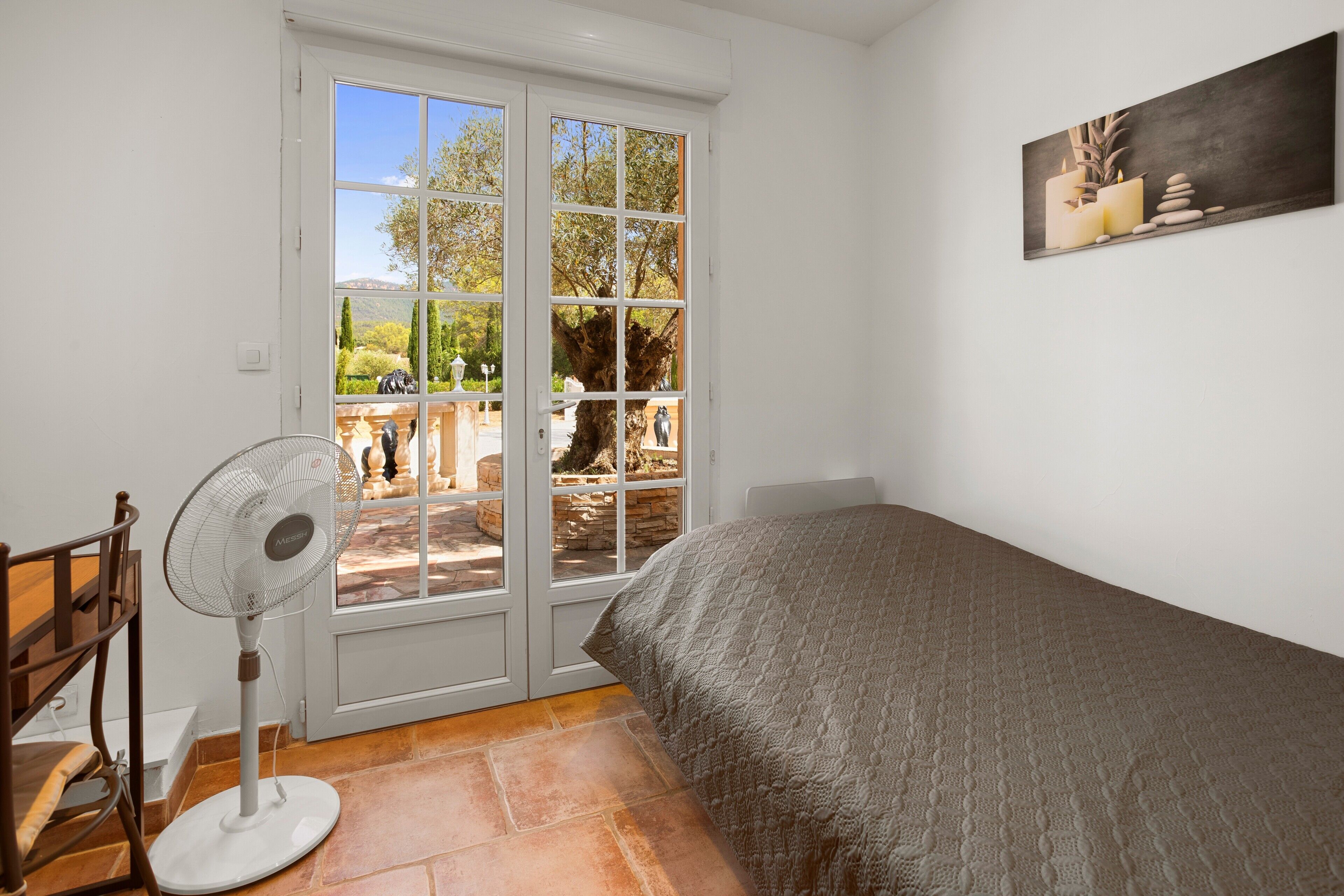 A bedroom with a bed, a standing fan, a wooden chair, and a view through a glass door.