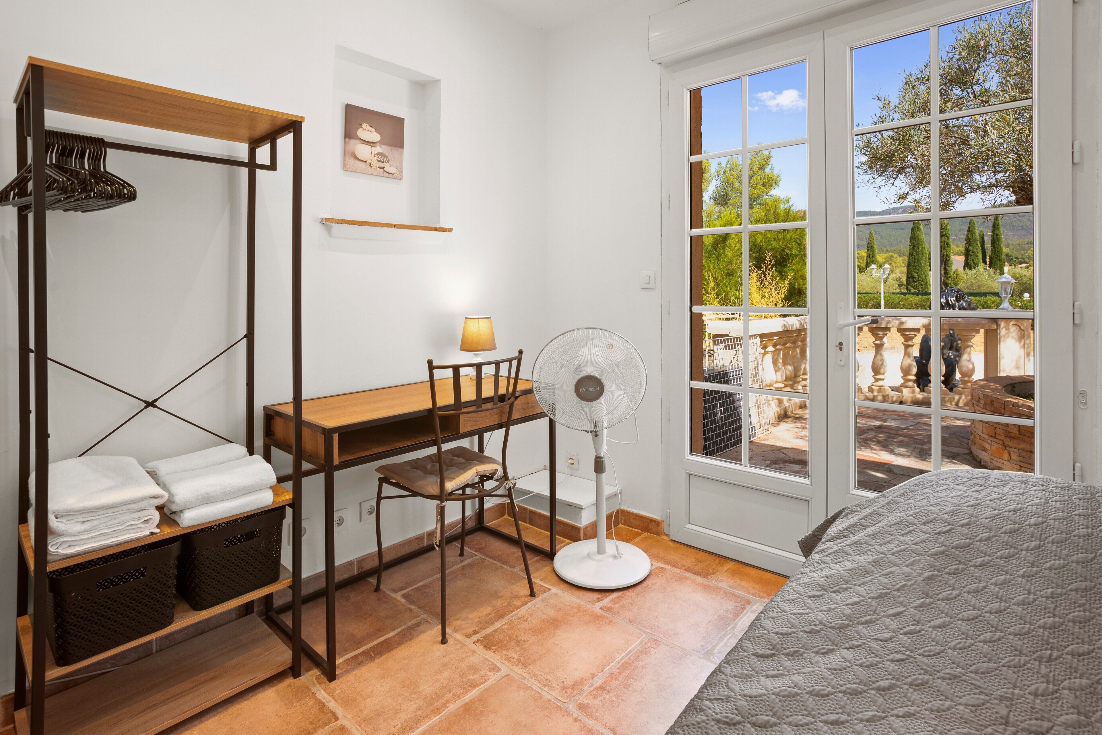 A bedroom with a bed, a desk, a chair, a fan, and a view of the outdoors.