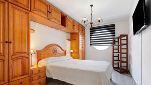 A bedroom with a bed, wooden wardrobe, bedside lamps, a TV, and a window with blinds. - Vacation Home 'Luha' with Private Terrace and Wi-Fi (Corralejo)
