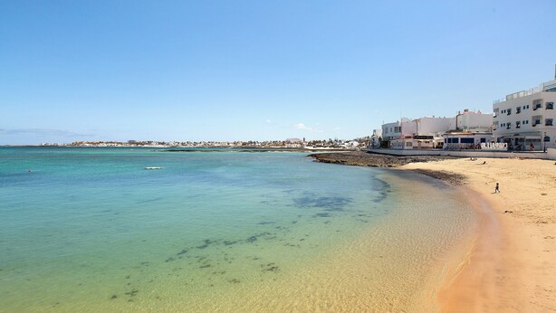 A beach with clear water, a sandy shore, and buildings in the background. - Vacation Home 'Luha' with Private Terrace and Wi-Fi (Corralejo)