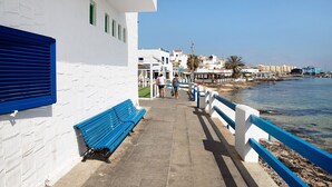 A coastal walkway with a blue bench, white buildings, and a clear view of the sea. - Vacation Home 'Luha' with Private Terrace and Wi-Fi (Corralejo)