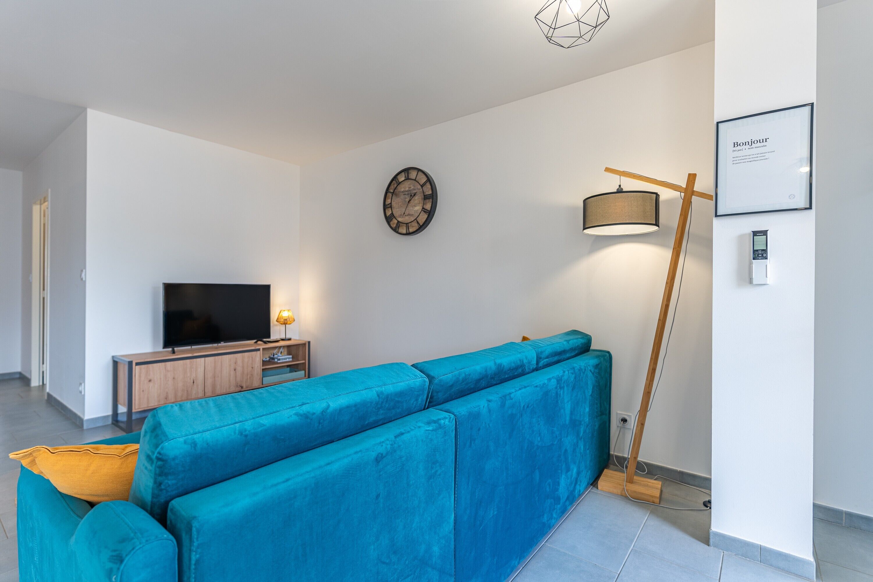 En moderne stue med en tealgrøn sofa, et fladskærms-tv, et træsidebord, et vægur, en gulvlampe og et indramet velkommensskilt.