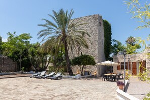 Une cour bordée de murs en pierre, agrémentée de palmiers, de chaises longues et de mobilier de jardin.