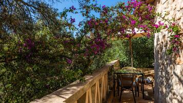 A stone balcony with a glass table and wicker chairs, surrounded by lush greenery and purple flowers.