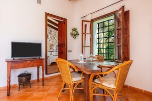 A dining area with a wooden table, wicker chairs, a television on a wooden stand, and a stone wall in the background.
