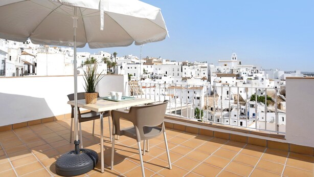 A terrace with a table, chairs, a white umbrella, and a view of white buildings.