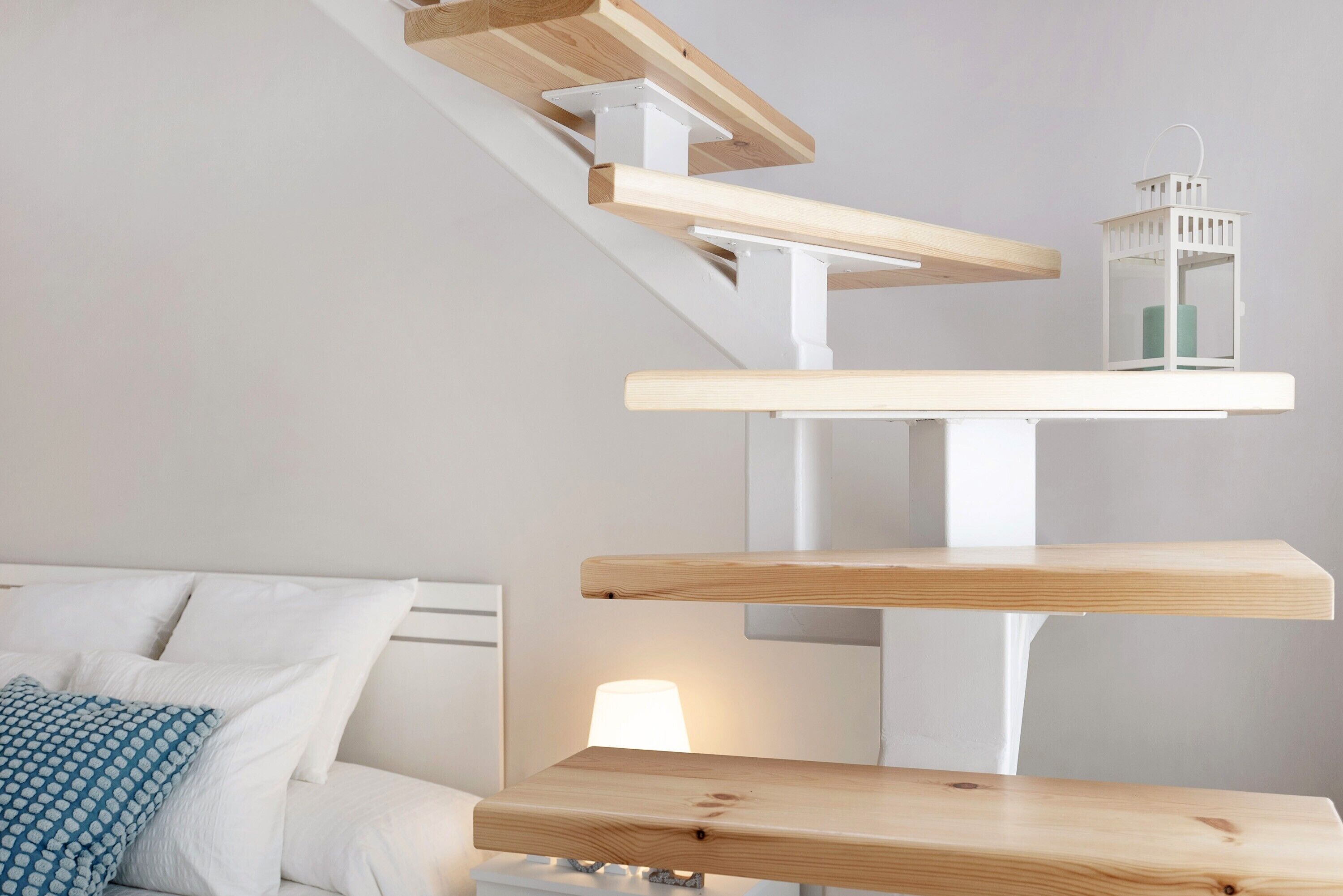 Un dormitorio con una cama, almohadas blancas, una almohada azul estampada, una mesita de noche con una lámpara y una pared blanca.