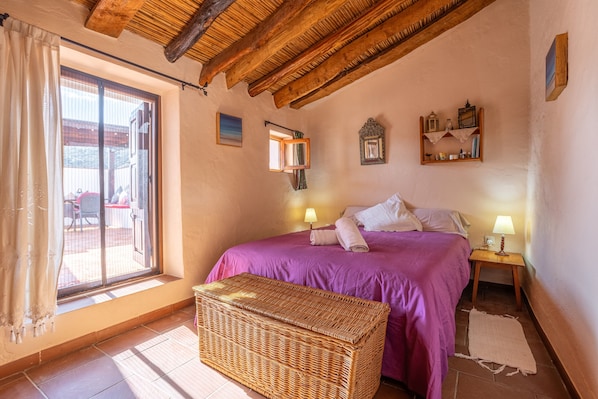 A bedroom with a bed, bedside lamps, a wicker bench, a window with curtains, and a wooden ceiling.