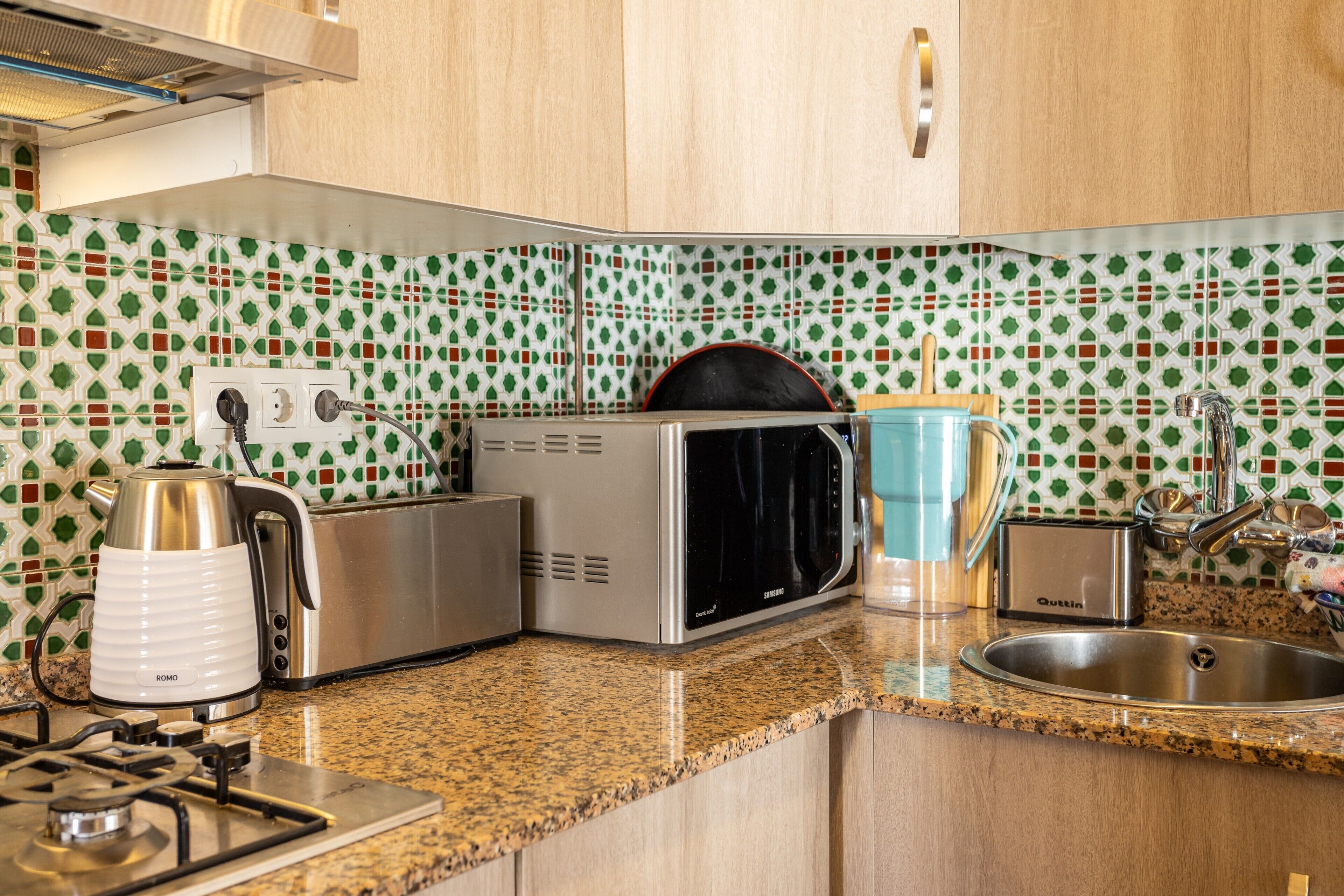 A kitchen with a microwave, toaster, kettle, sink, and a patterned backsplash.