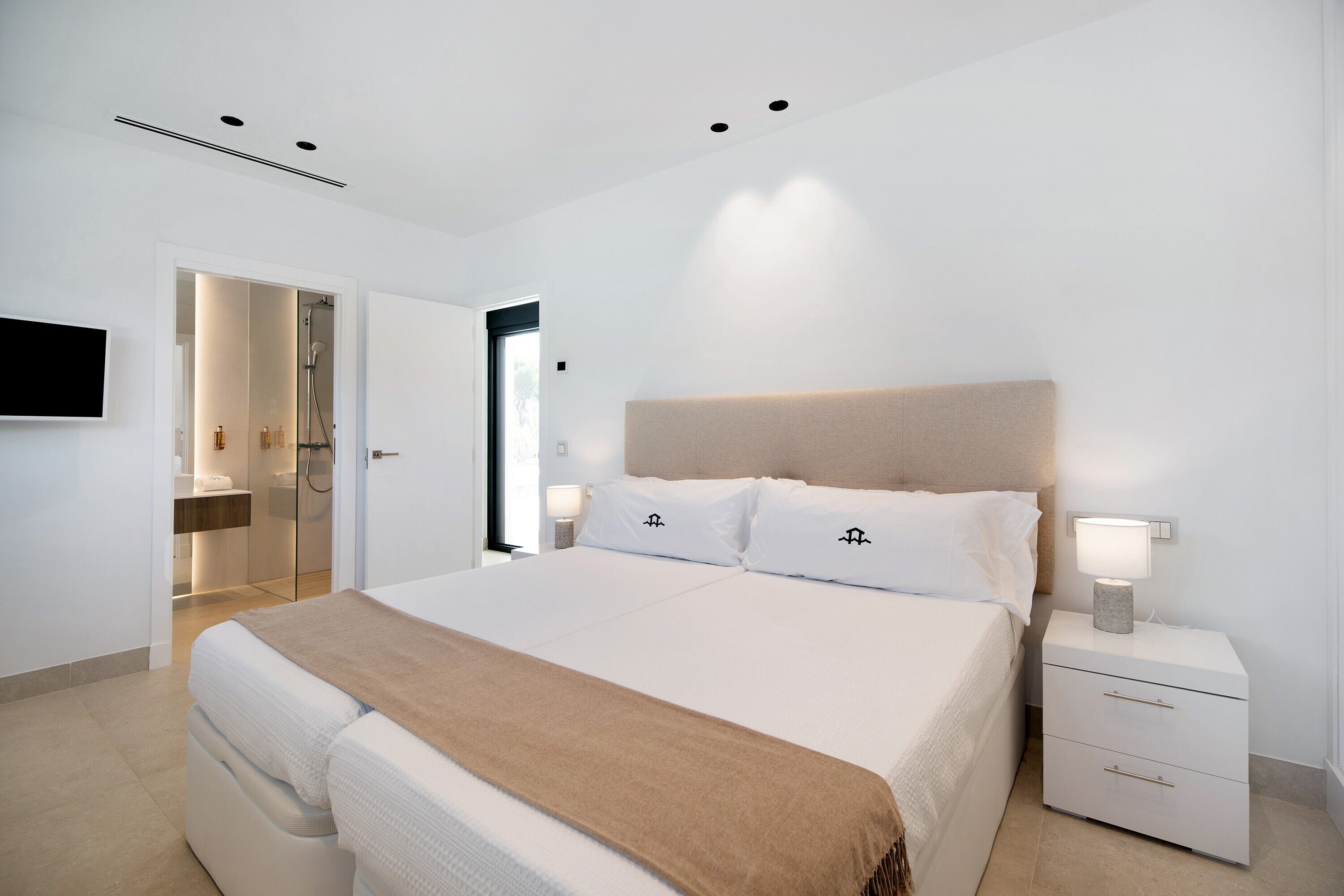 Een moderne slaapkamer met een groot bed, nachtkastjes, een televisie, en een zichtbare badkamer door een geopende deur.