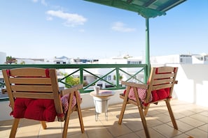 Deux chaises en bois avec des coussins rouges, sur un balcon doté d’une balustrade verte, une petite table avec un pichet blanc, et une vue dégagée sur des bâtiments blancs en arrière-plan.