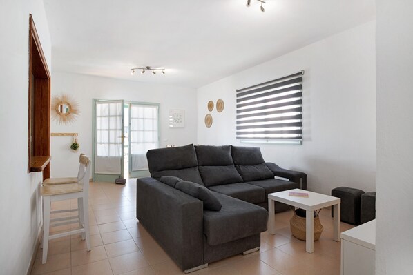 Un salon moderne avec un canapé d’angle, une petite table, un fauteuil et une fenêtre équipée de stores.