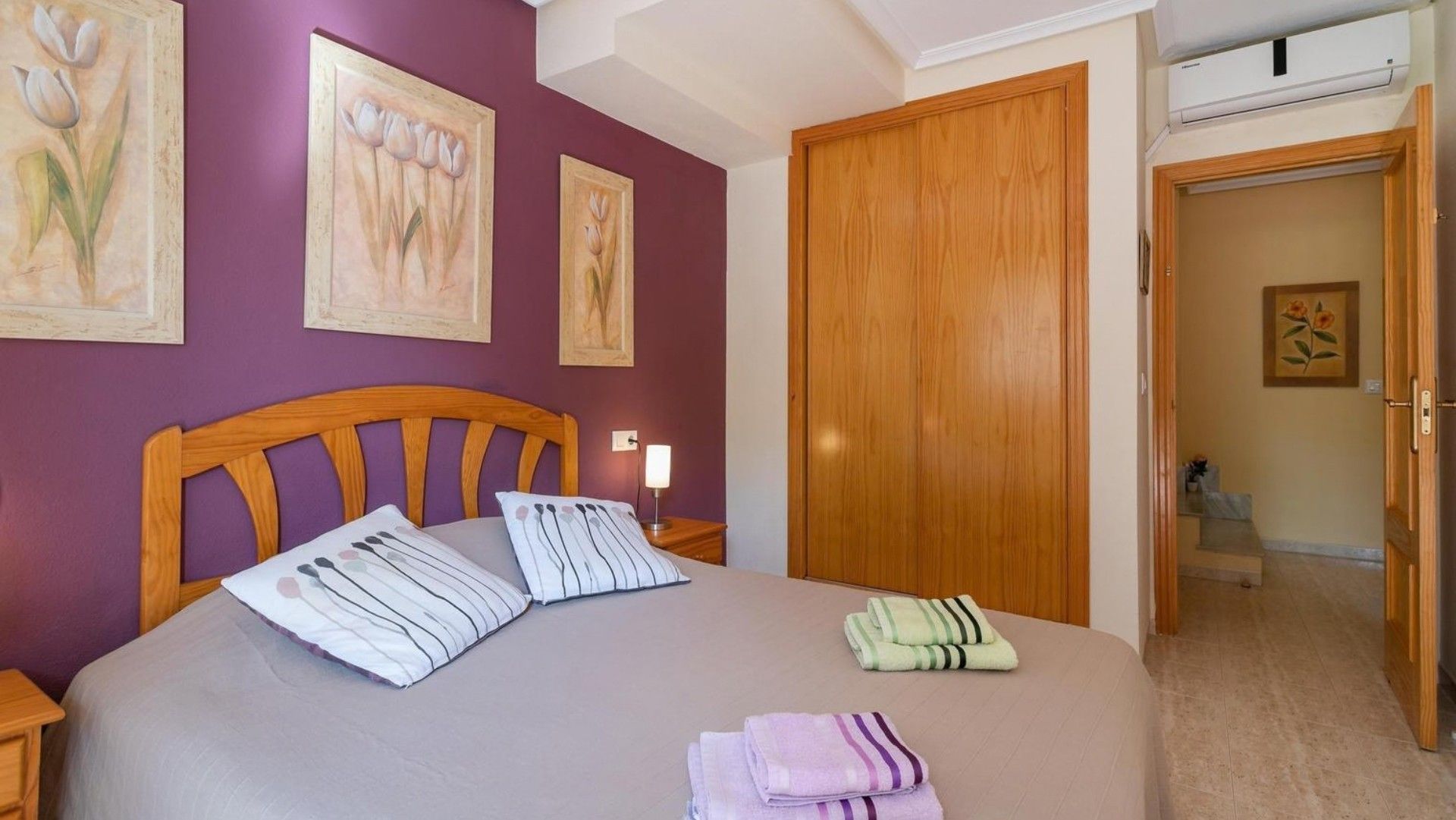 Un dormitorio con cama, almohadas, una mesita de noche, un armario de madera y una puerta que da a otra habitación.