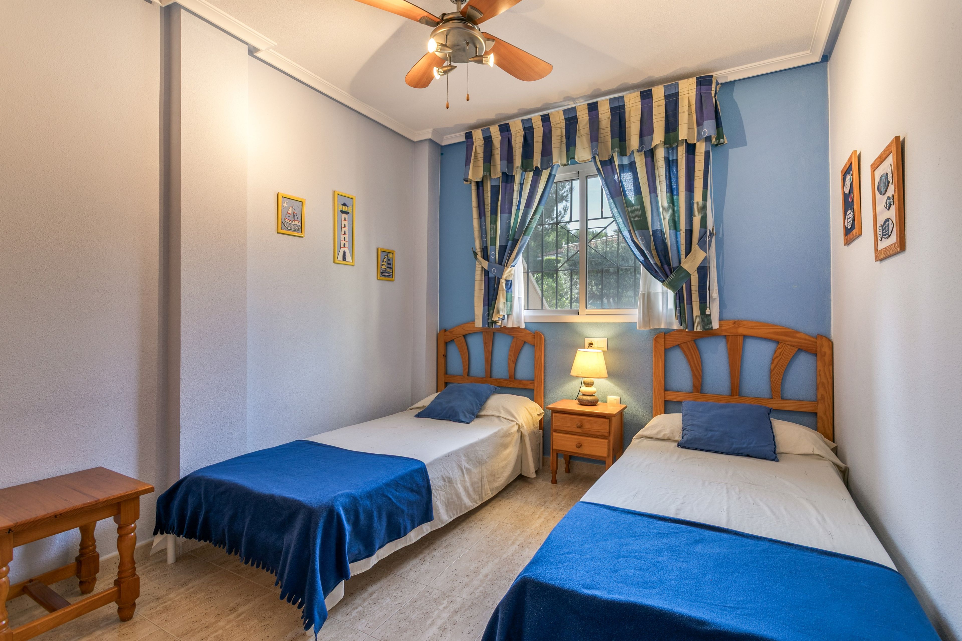 A bedroom with two single beds, a nightstand with a lamp, a wooden bench, and a ceiling fan.