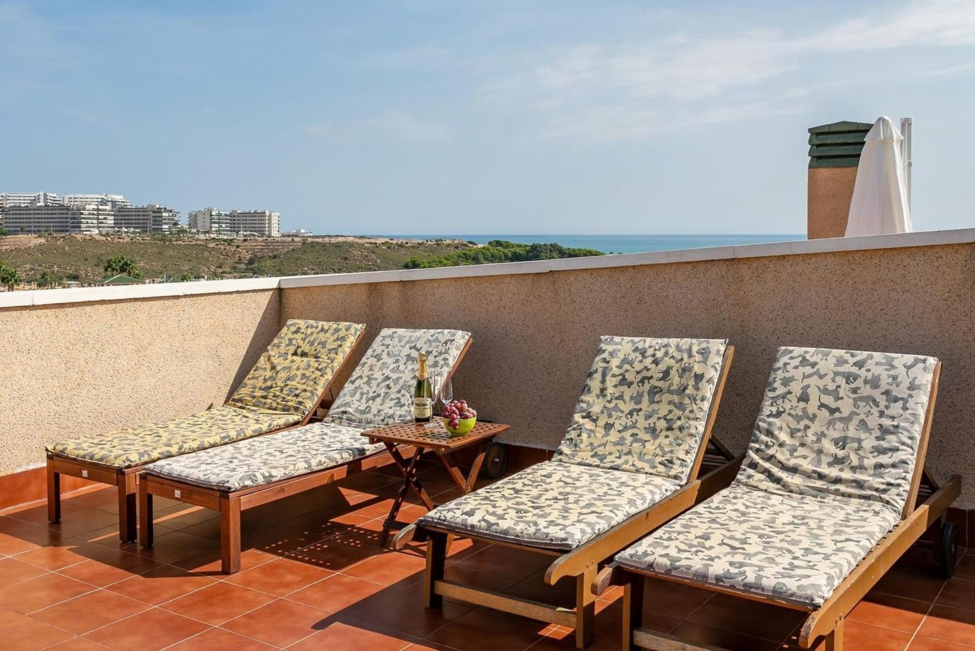 Una terraza en la azotea con cuatro sillones de madera y una mesa, con vistas a un paisaje costero y edificios a lo lejos.