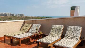 Una terraza en la azotea con cuatro sillones de madera y una mesa, con vistas a un paisaje costero y edificios a lo lejos.