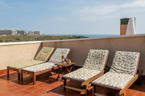 A rooftop terrace with four wooden lounge chairs and a table, overlooking a coastal landscape with buildings in the distance.