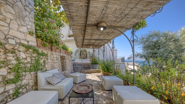 A stone wall with greenery, a covered seating area with cushions and a table, and a view of the sea. - Villa 'Blu La Patronale' with Sea View, Shared Pool and Wi-Fi (Castro)