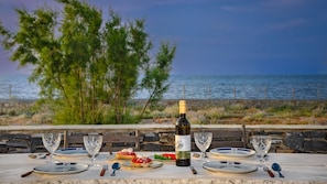 Mesa posta com pratos, talheres e uma garrafa de vinho, com vista para uma praia com árvores e o mar.