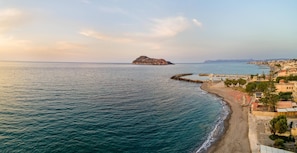 En kustvy med en klippig ö, en strand med parasoller och en pir som sträcker sig ut i havet.