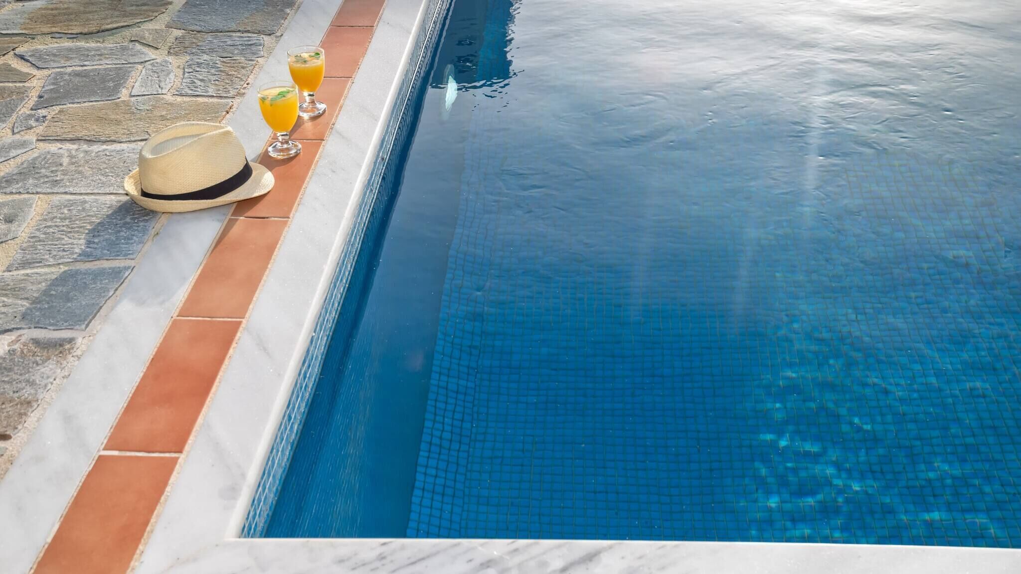 Une piscine avec un bord carrelé, un sol en pierre et deux verres de jus d’orange décorés de feuilles de menthe.
