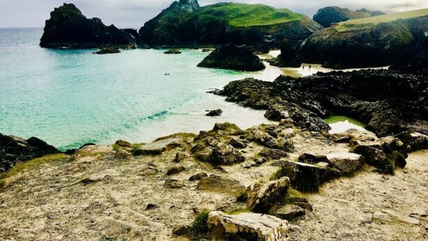 A rocky coastal landscape with clear turquoise waters and green hills in the background.