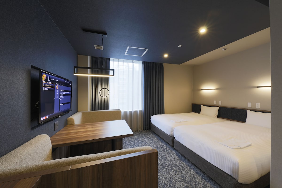 A modern hotel room with a flat-screen TV, a wooden desk, two single beds, and a large window with curtains.