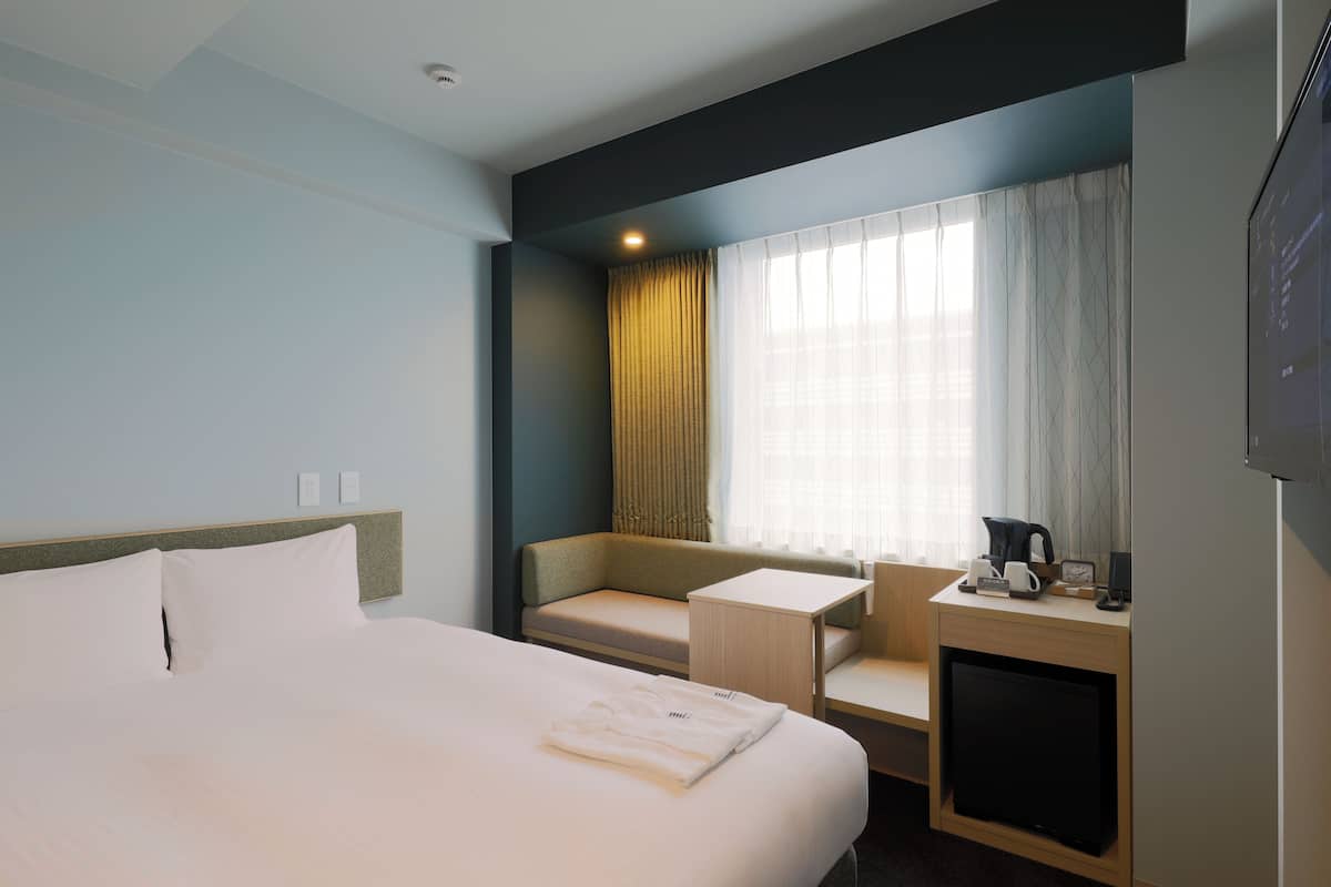 A hotel room with a bed, a seating area with a table, a TV, and a window with curtains.