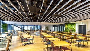 Une cafétéria spacieuse, dotée de longues tables et de chaises, de hauts plafonds avec des poutres apparentes et de grandes fenêtres laissant entrer la lumière naturelle.