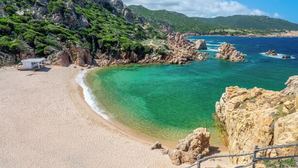 A beach with clear turquoise water, rocky cliffs, and a small building on the sand.