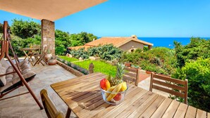 A wooden table with a fruit bowl, a stone pillar, and a view of the ocean.