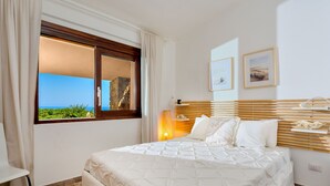 A bedroom with a large bed, a window with a view of the sea, and a nightstand with a lamp.
