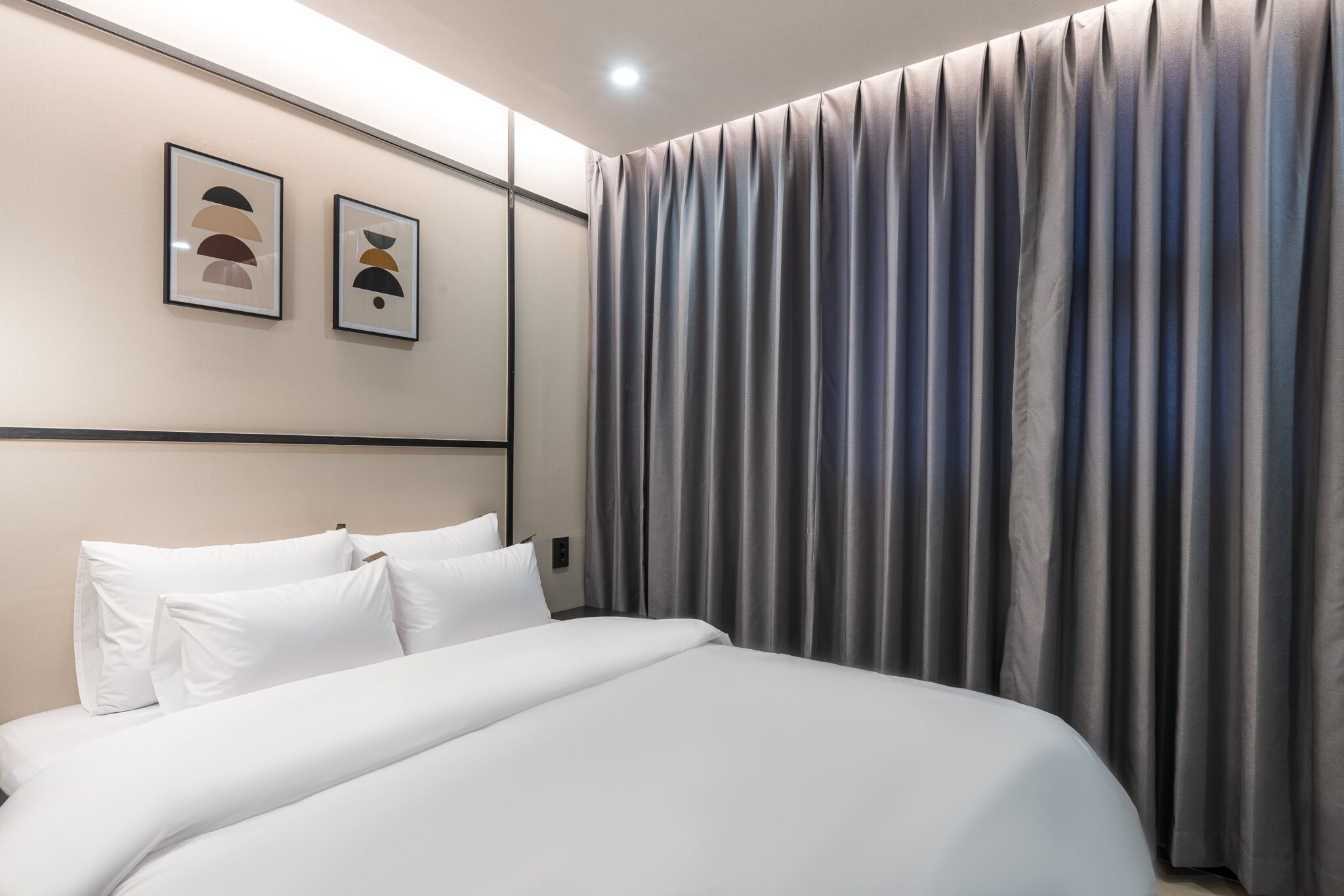A modern hotel room with a neatly made bed, a large window with curtains, and two framed artworks on the wall.