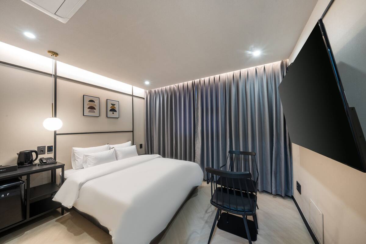A modern hotel room with a large bed, a desk with a lamp, a chair, a flat-screen TV, and curtains.