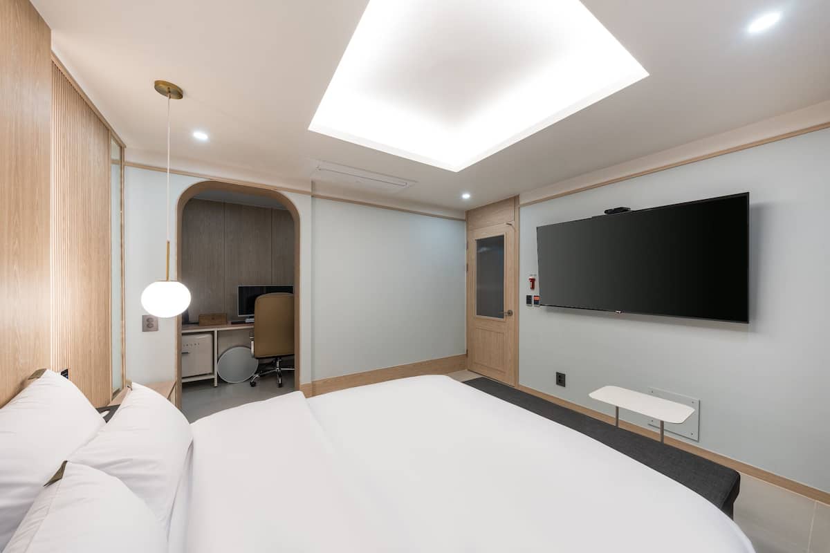 A modern hotel room with a large bed, a desk with a chair, a flat-screen TV mounted on the wall, and a skylight above the bed.