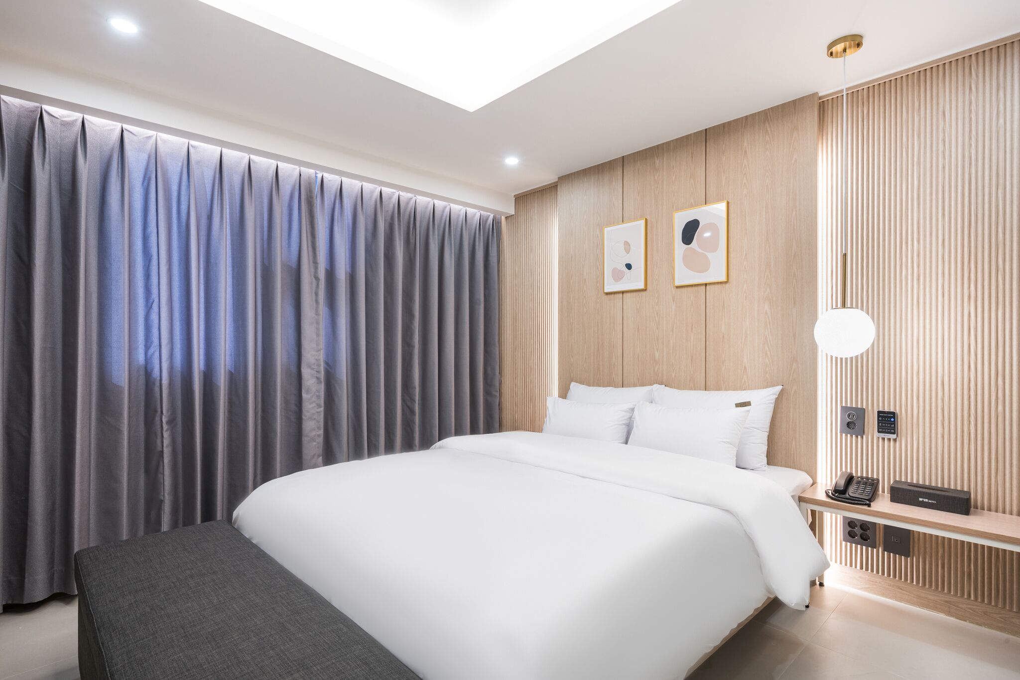 A modern hotel room with a large bed, a bedside table with a phone and a speaker, a wall-mounted light, and a wooden panel design.
