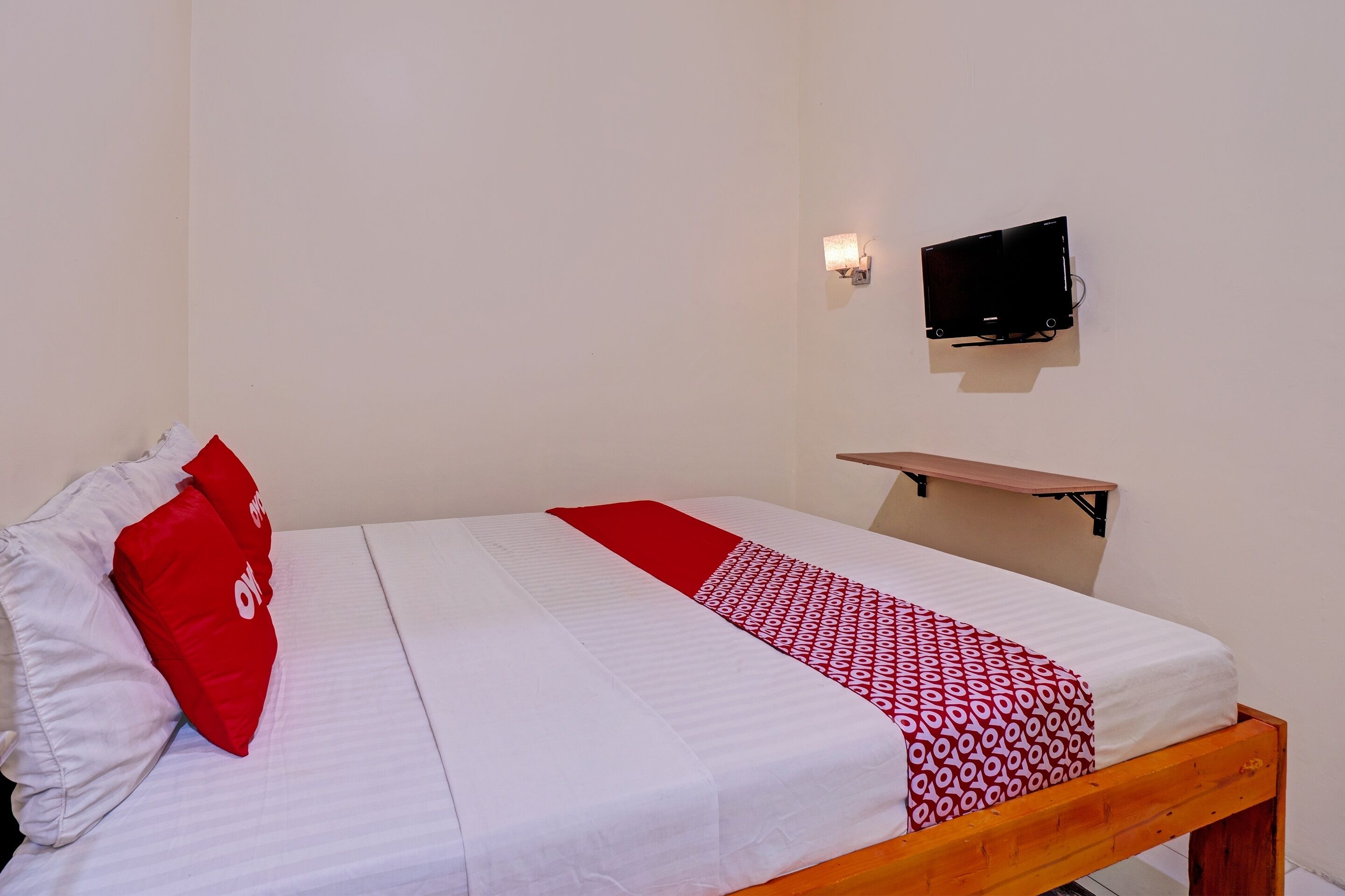Una cama bien hecha con ropa blanca y roja, una almohada roja y una mesita de noche de madera con un televisor encima.