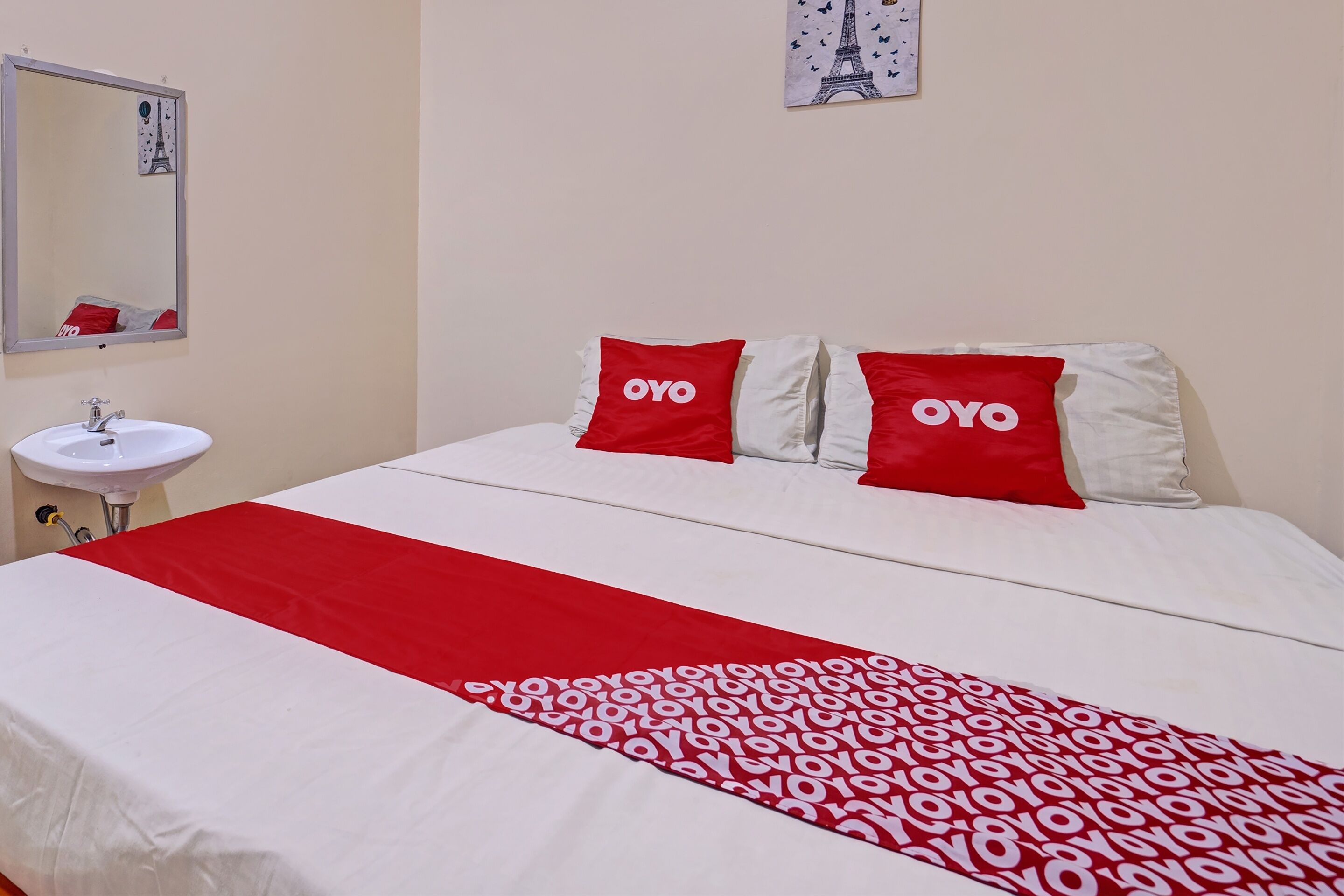 A hotel room with a bed, two red pillows with "OYO" branding, a white and red bedspread, a white sink, and a mirror with an Eiffel Tower image.