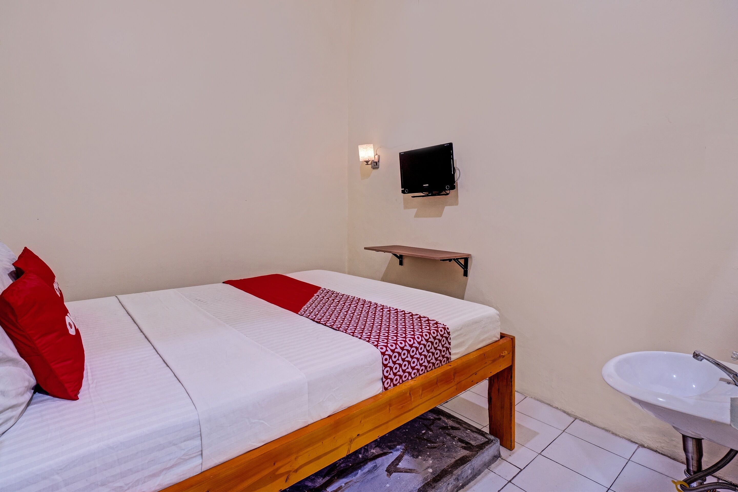 Una cama individual con estructura de madera, ropa de cama blanca y almohada roja. Un estante con un televisor está montado en la pared sobre la cama. Se ve un lavabo blanco a la derecha.
