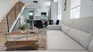 A modern living room with a grey sofa, a wooden coffee table, a staircase, and a kitchen in the background. - MODERN 4BR TOWNHOME BUILT 2023 w/ 2-CAR GARAGE WITH EV CHARGER & 2 BALCONIES (p63) (Los Angeles)