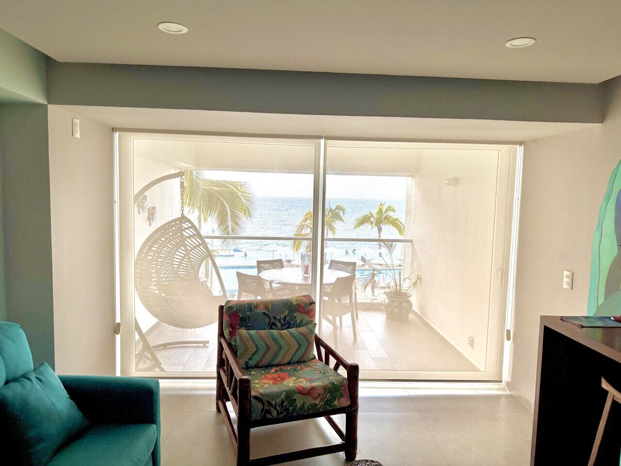 Ein Zimmer mit einer großen Schiebeglastür, die auf einen Balkon mit Pool und Palmen führt. Ein Stuhl mit Blumenkissen und ein türkisfarbenes Sofa sind ebenfalls zu sehen.