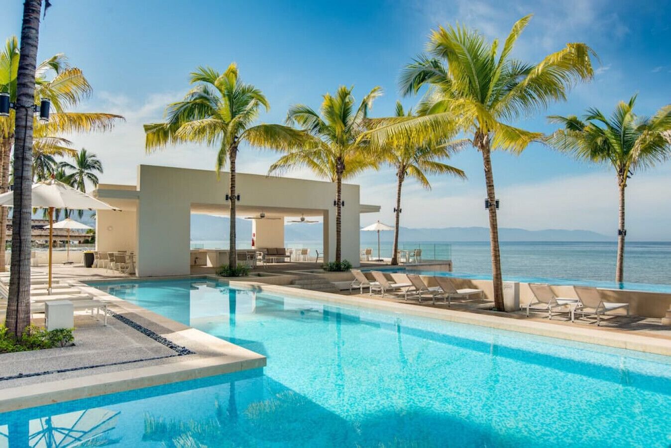 Un espace aménagé au bord de la piscine, avec des chaises longues et des parasols, des palmiers et une vue sur l’océan.