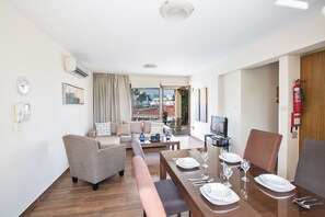 A modern living room with a sofa, armchairs, a dining table set for four, and a television. - Ayia Napa Holiday Apartment Na101 (Ayia Napa)