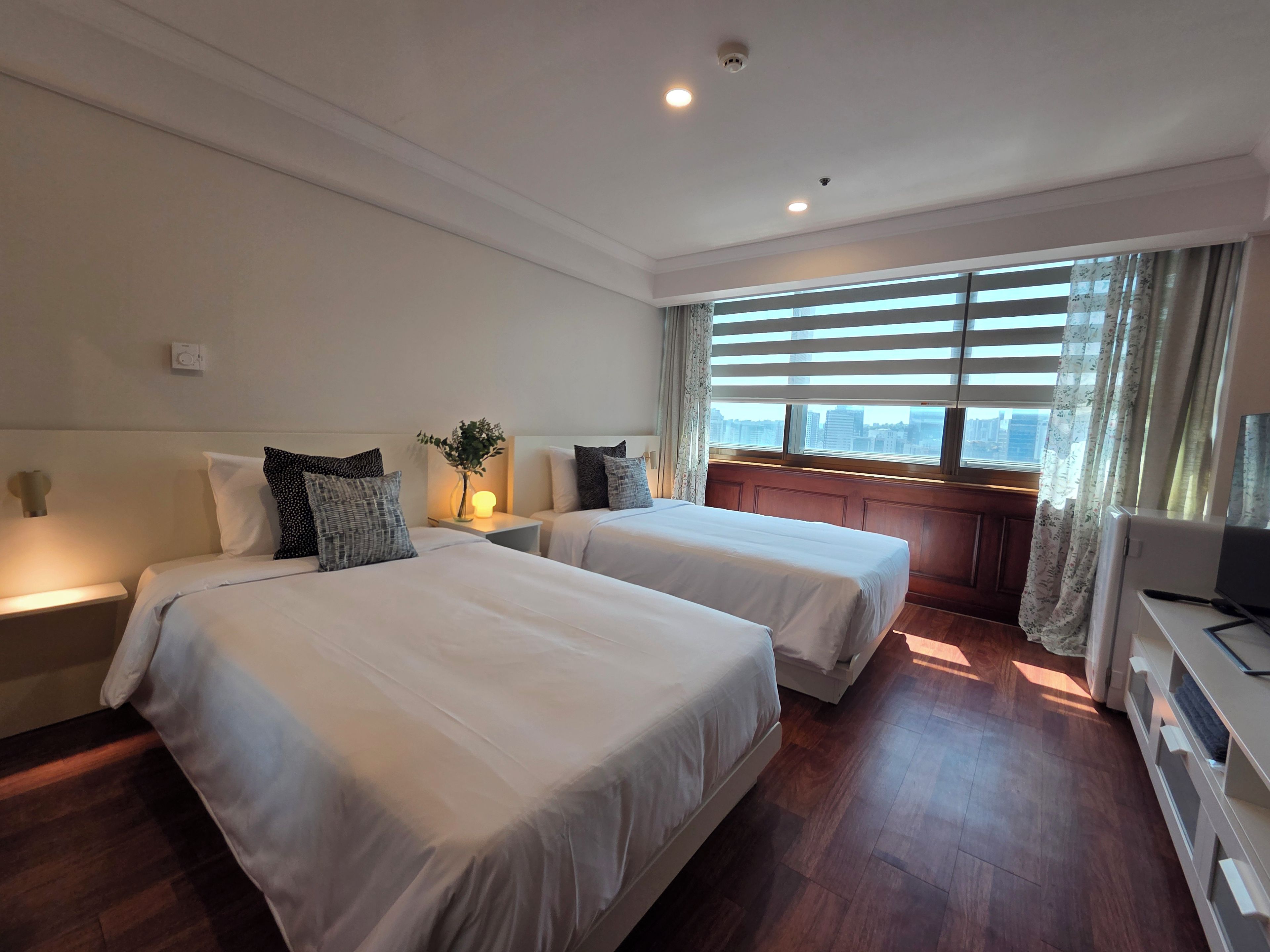 A hotel room with two beds, a nightstand with a lamp, a window with blinds, and a city view.