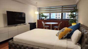 Habitación de hotel moderna con una cama grande, un televisor de pantalla plana, una mesita con sillas y una ventana con persianas.