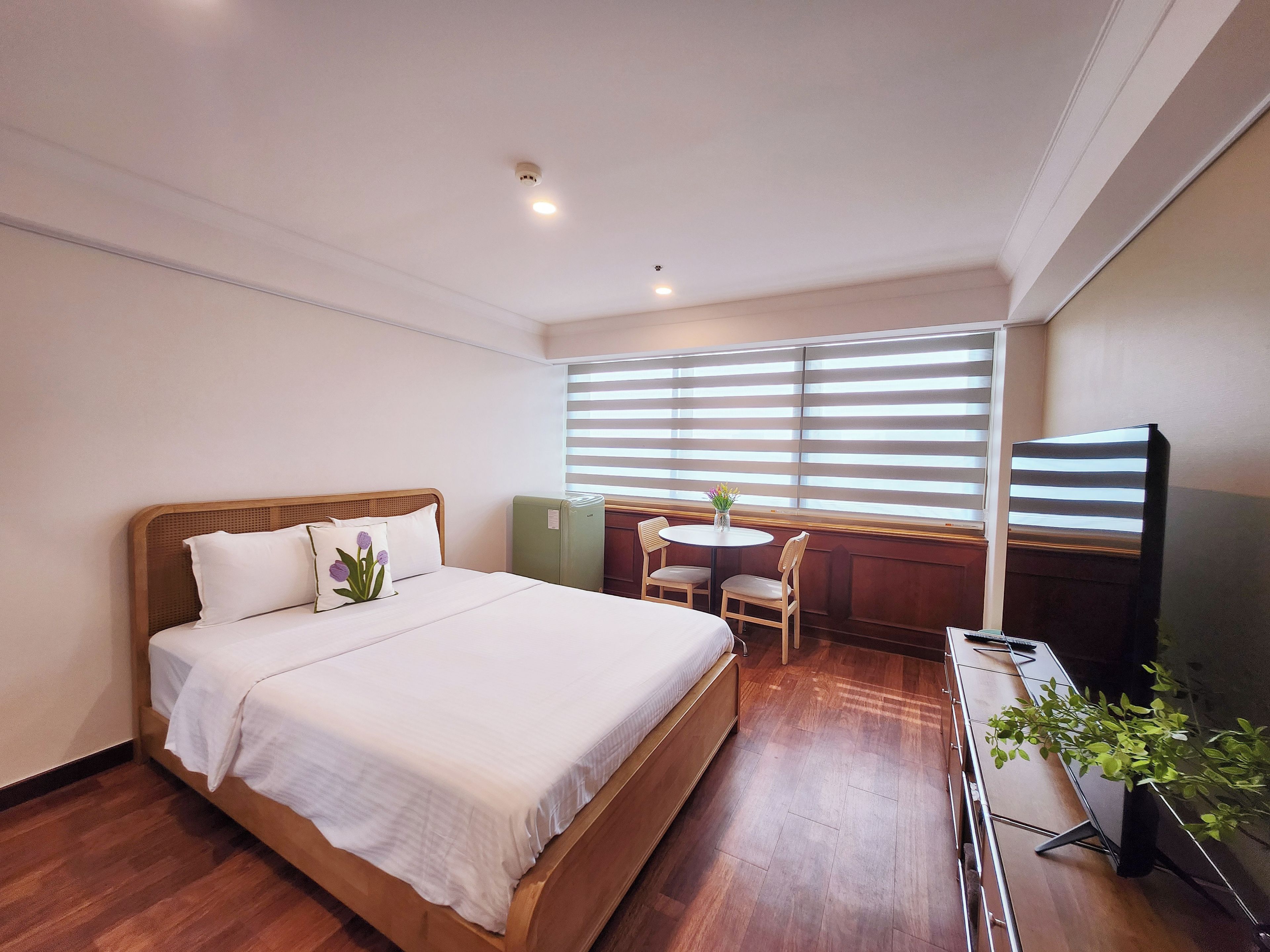 A hotel room with a wooden bed, a small table with chairs, a television, and a window with blinds.