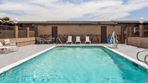 A rectangular swimming pool with clear blue water, surrounded by a low stone wall. There are lounge chairs and a small table on the poolside. - Tri City Inn (Victorville)